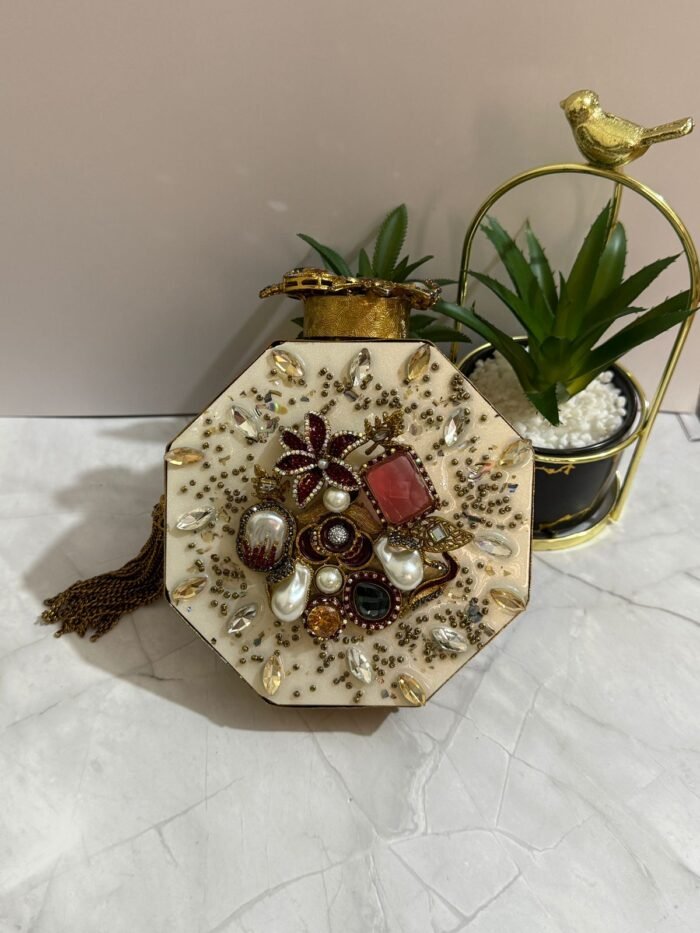 A luxurious octagonal clutch with gold accents, jewel-toned crystals, pearls, and a tassel detail, placed alongside decorative plants.
