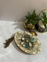 A luxurious oval clutch with green gemstones, pearls, and gold detailing, finished with a tassel, placed alongside decorative plants.