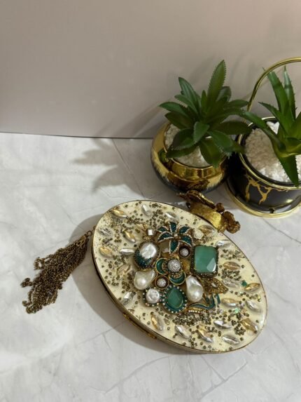 A luxurious oval clutch with green gemstones, pearls, and gold detailing, finished with a tassel, placed alongside decorative plants.