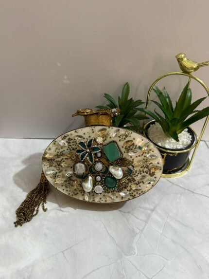 A luxurious oval clutch with green gemstones, pearls, and gold detailing, finished with a tassel, placed alongside decorative plants.