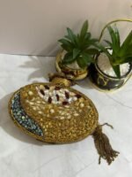 A luxurious oval clutch with green gemstones, pearls, and gold detailing, finished with a tassel, placed alongside decorative plants.