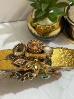 A luxurious oval clutch with green gemstones, pearls, and gold detailing, finished with a tassel, placed alongside decorative plants.