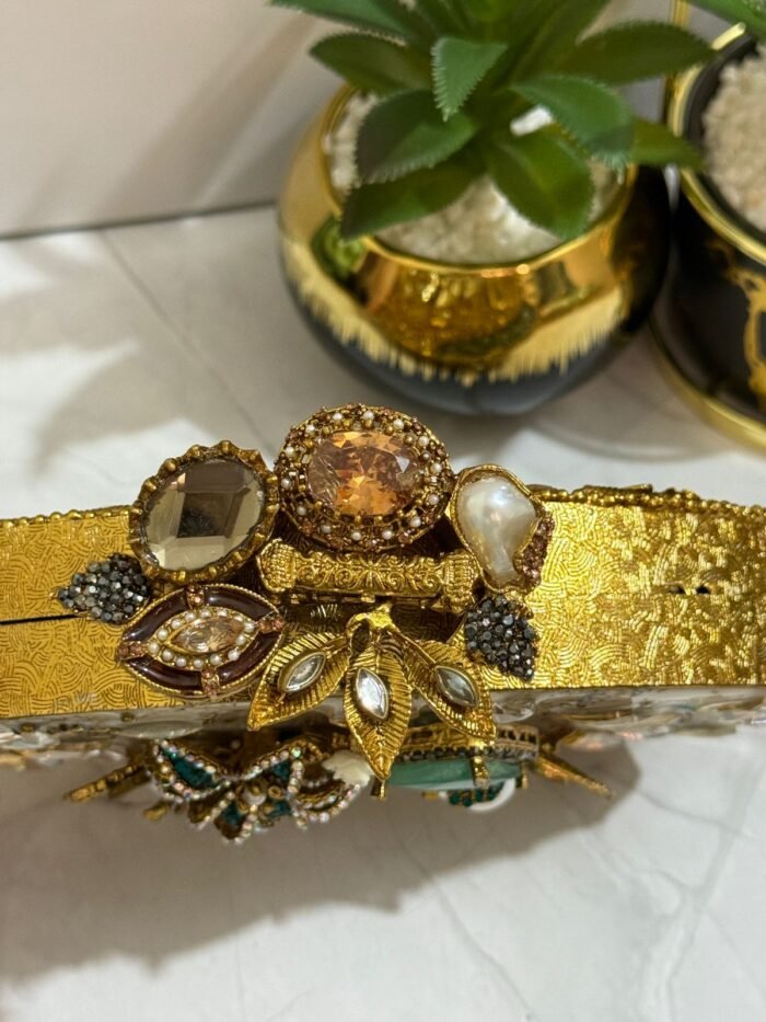 A luxurious oval clutch with green gemstones, pearls, and gold detailing, finished with a tassel, placed alongside decorative plants.