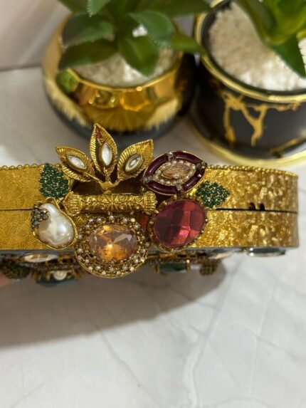 A luxurious octagonal clutch with purple and gold embellishments, pearls, and crystals, finished with a gold tassel, placed next to decorative plants.