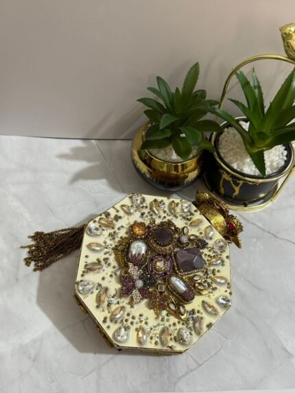 A luxurious octagonal clutch with purple and gold embellishments, pearls, and crystals, finished with a gold tassel, placed next to decorative plants.