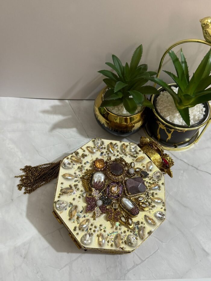 A luxurious octagonal clutch with purple and gold embellishments, pearls, and crystals, finished with a gold tassel, placed next to decorative plants.