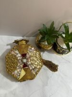 A luxurious octagonal clutch with purple and gold embellishments, pearls, and crystals, finished with a gold tassel, placed next to decorative plants.