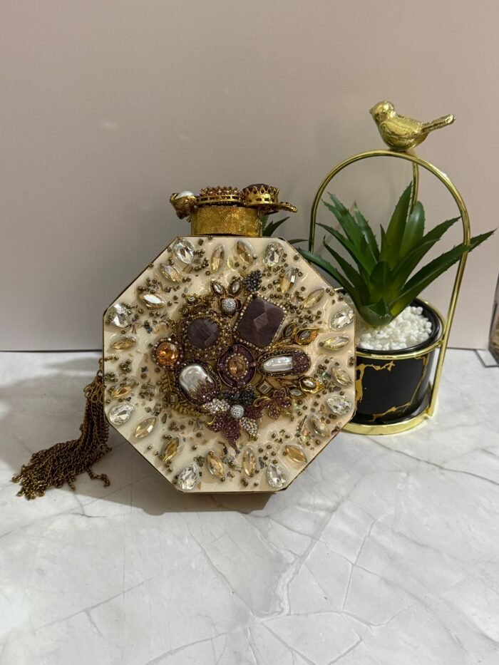 A luxurious octagonal clutch with purple and gold embellishments, pearls, and crystals, finished with a gold tassel, placed next to decorative plants.