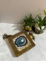 A square clutch with lapis lazuli, pearl, and crystal embellishments, surrounded by gold-tone wirework, with a gold tassel, placed next to decorative plants.