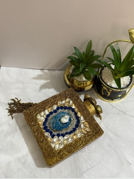 A square clutch with lapis lazuli, pearl, and crystal embellishments, surrounded by gold-tone wirework, with a gold tassel, placed next to decorative plants.