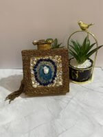 A square clutch with lapis lazuli, pearl, and crystal embellishments, surrounded by gold-tone wirework, with a gold tassel, placed next to decorative plants.
