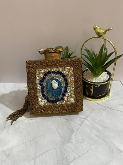 A square clutch with lapis lazuli, pearl, and crystal embellishments, surrounded by gold-tone wirework, with a gold tassel, placed next to decorative plants.