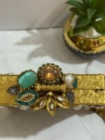 A square clutch with lapis lazuli, pearl, and crystal embellishments, surrounded by gold-tone wirework, with a gold tassel, placed next to decorative plants.
