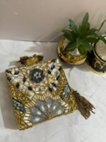 A square clutch with lapis lazuli, pearl, and crystal embellishments, surrounded by gold-tone wirework, with a gold tassel, placed next to decorative plants.