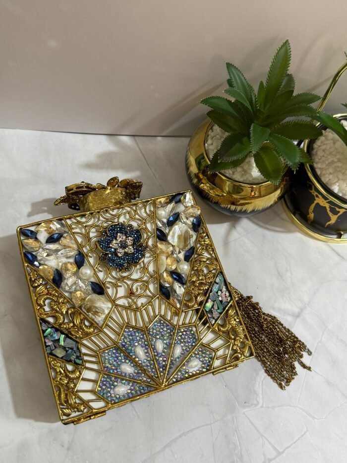 A square clutch with lapis lazuli, pearl, and crystal embellishments, surrounded by gold-tone wirework, with a gold tassel, placed next to decorative plants.