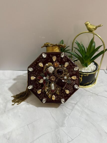 An octagonal clutch with red and gold gemstone embellishments, crystals, pearls, and a gold tassel, placed beside decorative plants.