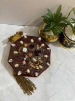 An octagonal clutch with red and gold gemstone embellishments, crystals, pearls, and a gold tassel, placed beside decorative plants.