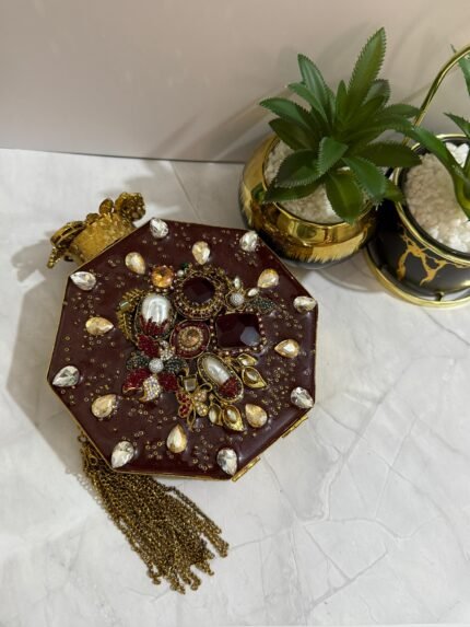 An octagonal clutch with red and gold gemstone embellishments, crystals, pearls, and a gold tassel, placed beside decorative plants.
