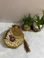 An octagonal clutch with red and gold gemstone embellishments, crystals, pearls, and a gold tassel, placed beside decorative plants.