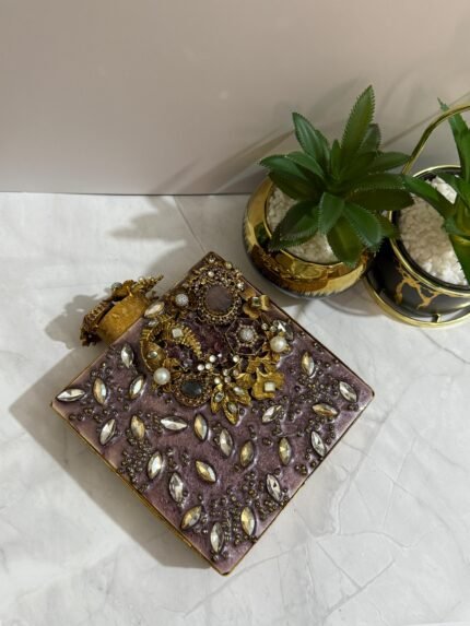 A square clutch with crystal and gold floral embellishments, featuring pearls and clear crystals, placed next to decorative plants.