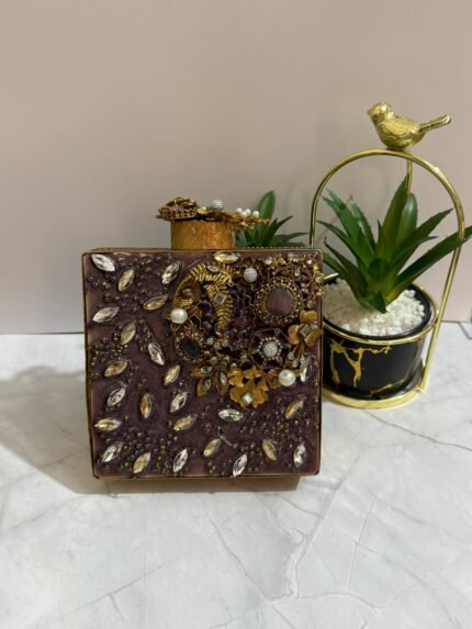 A square clutch with crystal and gold floral embellishments, featuring pearls and clear crystals, placed next to decorative plants.