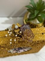 A square clutch with crystal and gold floral embellishments, featuring pearls and clear crystals, placed next to decorative plants.