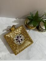 A square clutch with crystal and gold floral embellishments, featuring pearls and clear crystals, placed next to decorative plants.