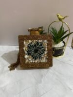 A square clutch with a green beaded floral design, crystal embellishments, and a tassel, placed next to decorative plants.
