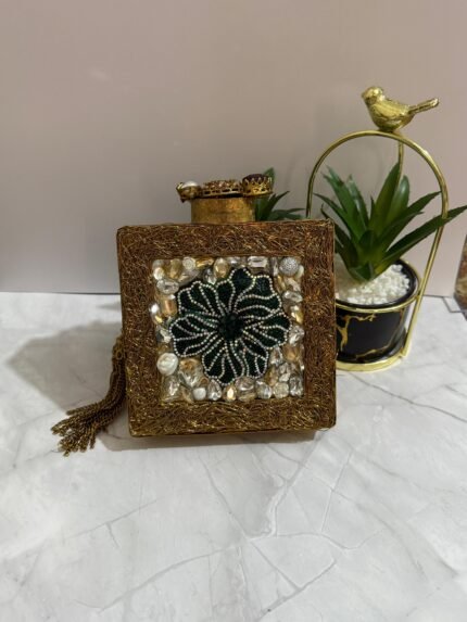 A square clutch with a green beaded floral design, crystal embellishments, and a tassel, placed next to decorative plants.