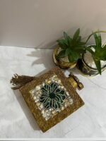A square clutch with a green beaded floral design, crystal embellishments, and a tassel, placed next to decorative plants.