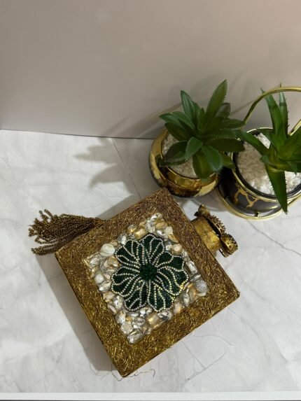 A square clutch with a green beaded floral design, crystal embellishments, and a tassel, placed next to decorative plants.