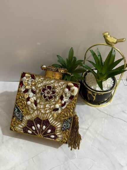 A gold clutch with a crystal and pearl floral design, placed next to decorative plants, with an attached tassel detail.