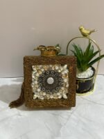 A gold clutch with a crystal and pearl floral design, placed next to decorative plants, with an attached tassel detail.