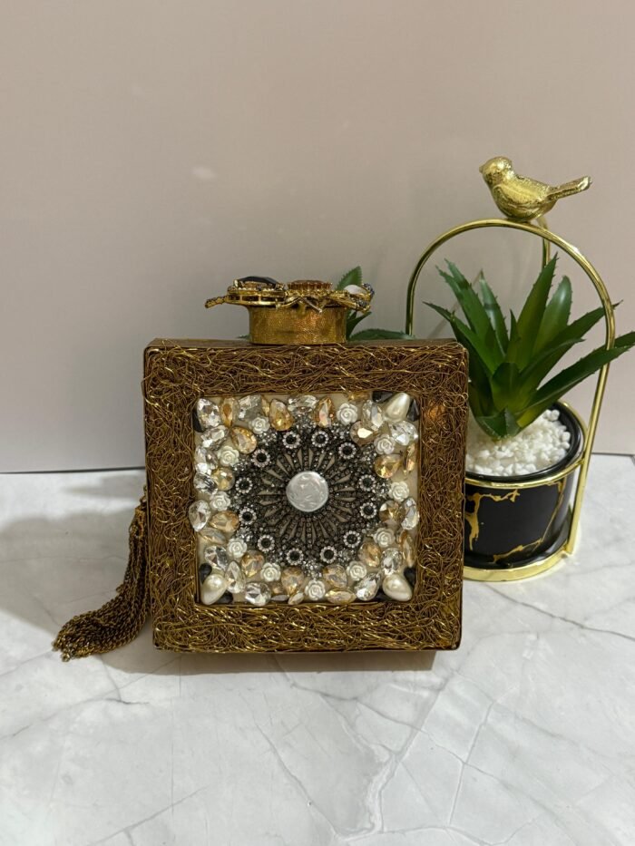 A gold clutch with a crystal and pearl floral design, placed next to decorative plants, with an attached tassel detail.