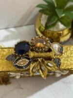 A gold clutch with a crystal and pearl floral design, placed next to decorative plants, with an attached tassel detail.