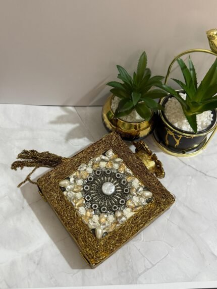 A gold clutch with a crystal and pearl floral design, placed next to decorative plants, with an attached tassel detail.