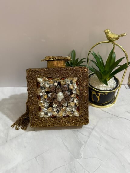 A gold clutch with a crystal and pearl flower embellishment, placed next to decorative plants, with a gold tassel attached.