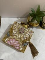 A gold clutch with a crystal and pearl flower embellishment, placed next to decorative plants, with a gold tassel attached.