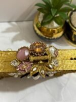 A gold clutch with a crystal and pearl flower embellishment, placed next to decorative plants, with a gold tassel attached.