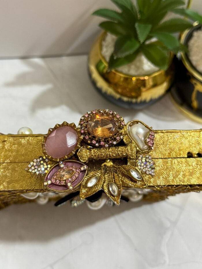 A gold clutch with a crystal and pearl flower embellishment, placed next to decorative plants, with a gold tassel attached.