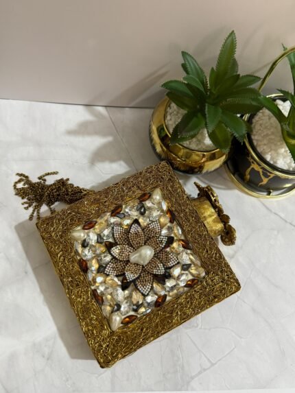 A gold clutch with a crystal and pearl flower embellishment, placed next to decorative plants, with a gold tassel attached.
