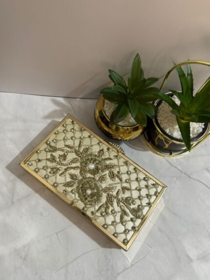 A cream-colored beaded clutch with floral design and pearl accents, placed next to decorative plants.