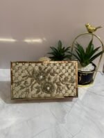 A cream-colored beaded clutch with floral design and pearl accents, placed next to decorative plants.