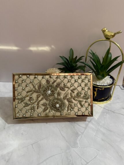 A cream-colored beaded clutch with floral design and pearl accents, placed next to decorative plants.
